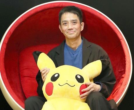 Image Description: The creator of Pokémon, Satoshi Tajiri picture with a plush Pikachu. He is sitting in an egg chair that is red on the inside. He is wearing a black blazer with a blue top and black trousers. He is holding a large pikachu plush toy between his legs.