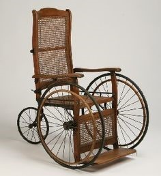 A drawing of an 19th century wooden wheelchair. The two front wheels are large, with one small back wheel. There is also a foot rest