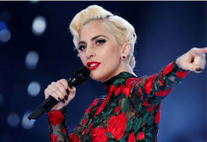 Lady Gaga is shown from the chest up holding a microphone to her mouth and pointing off to the right of the image. Her her is blonde and in a bun. She is wearing lace covered in bright red roses and green vines.