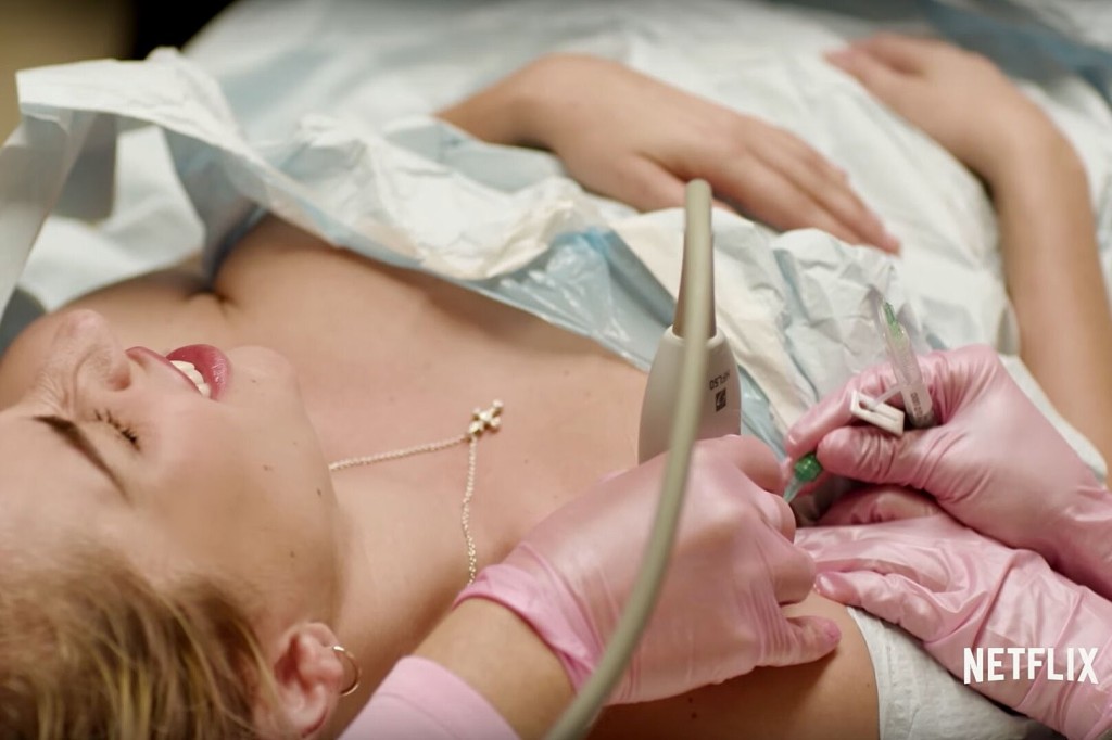 Lady Gaga is shown lying under a plastic sheet, wincing in pain. There are medically gloved hands - one holding an ultrasound machine and two administering a needle into her shoulder.