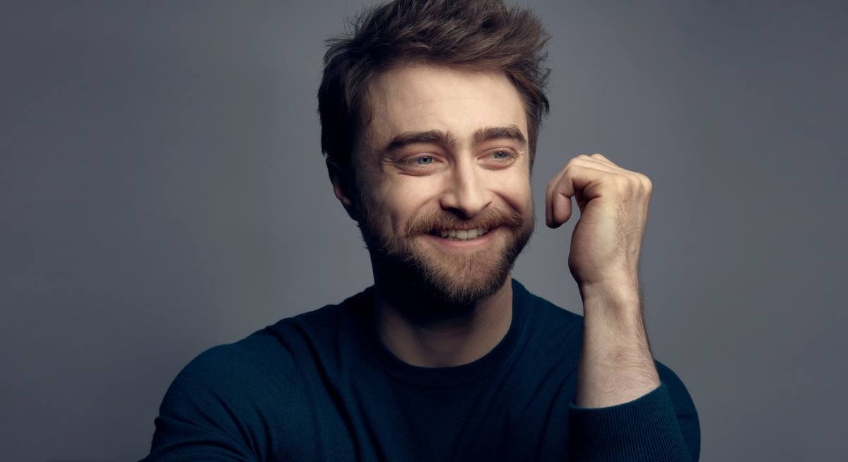 A close up photograph of Daniel Radcliffe's face. He is smiling and looking off to the right hand side. He has a neat short beard and his short brown hair is styled to stand on end. He is wearing a blue crew-neck, long sleeve top. His left arm is held up by his face, but not touching it, his hand curled into an open fist.