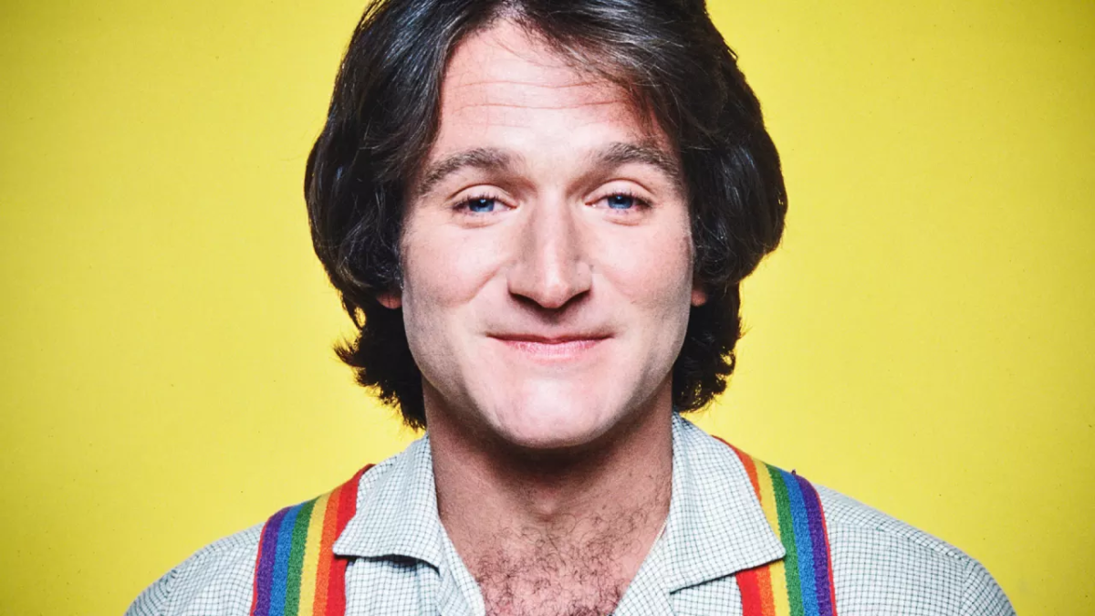 A young Robin Williams smiling with a yellow background. he is wearing a white shirt with rainbow braces.