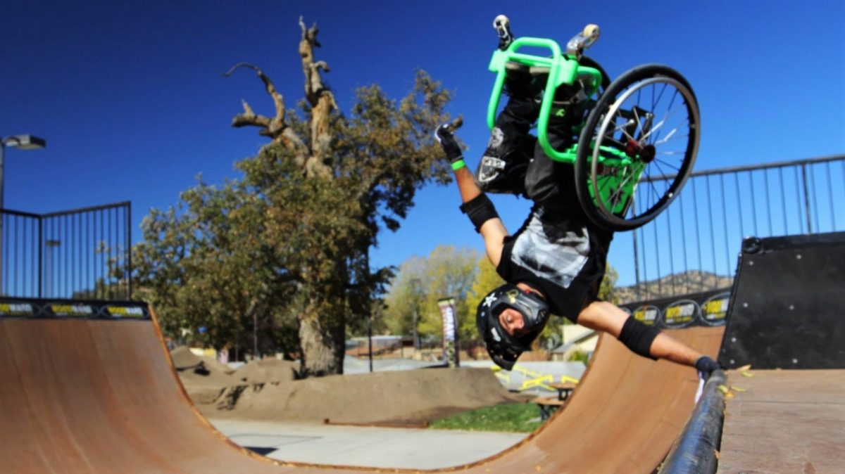 Aaron Fotheringham on a green wheelchair, wearing a helmet. He is upside down in the air with his hand on the edge of a half pipe.