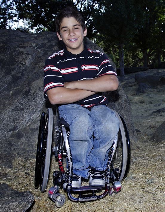 A young Aaron Fotheringham sitting in his wheelchair smiling with his arms crossed. He is wearing a navy blue, red and white stripped polo shirt with blue denim jeans.
In the background behind him is a large rock and some trees.