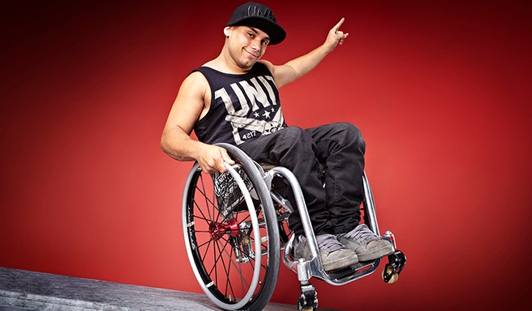 Aaron Fotheringham in his wheelchair on one back wheel doing a wheelie, smiling and his arm to the side and his hand with his pointing finger pointing up. He is wearing black denim jeans, a black vest top with decorative imaging on it, dirty white trainers and a black cap on his head.
The background is red.