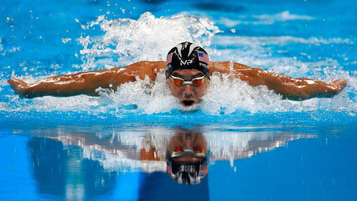 Michael Phelps swimming through the water in a pool towards the camera. He is wearing a swimming cap and goggles.