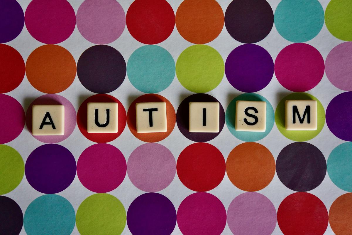 Autism spelt out in scrabble tiles on a spotty background. The spots are different colours and the scrabble tiles are across the middle row, inside one dot each.