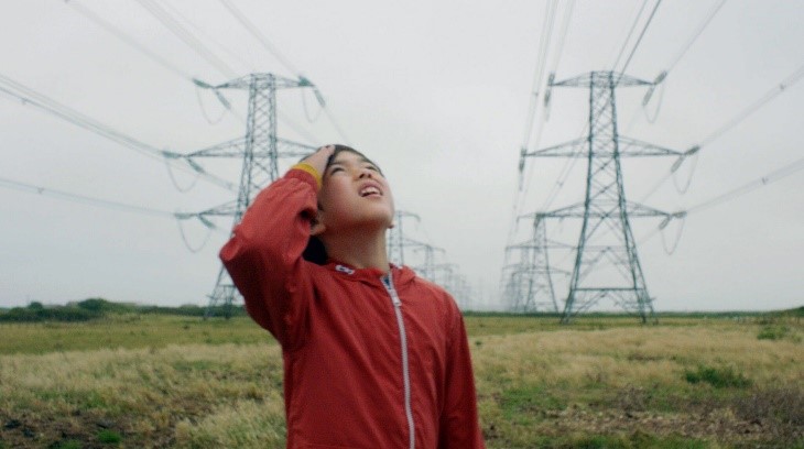 Screenshot from the film the reason I jump. Young boy is looking up towards the sky. He is wearing a red raincoat. In the background there are pylons.