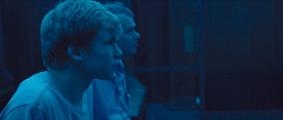 A screenshot from the film the reason I jump. Everything is in tones of blue. You can see a young man in the forefront looking to the right. A behind him another man looking the same way.