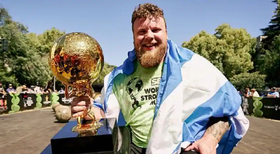 Tom Stolotman holding a gol trophy and smiling at the camera. He is a white man with short curly hair and a beard. He has a scottish flag around his shoulders