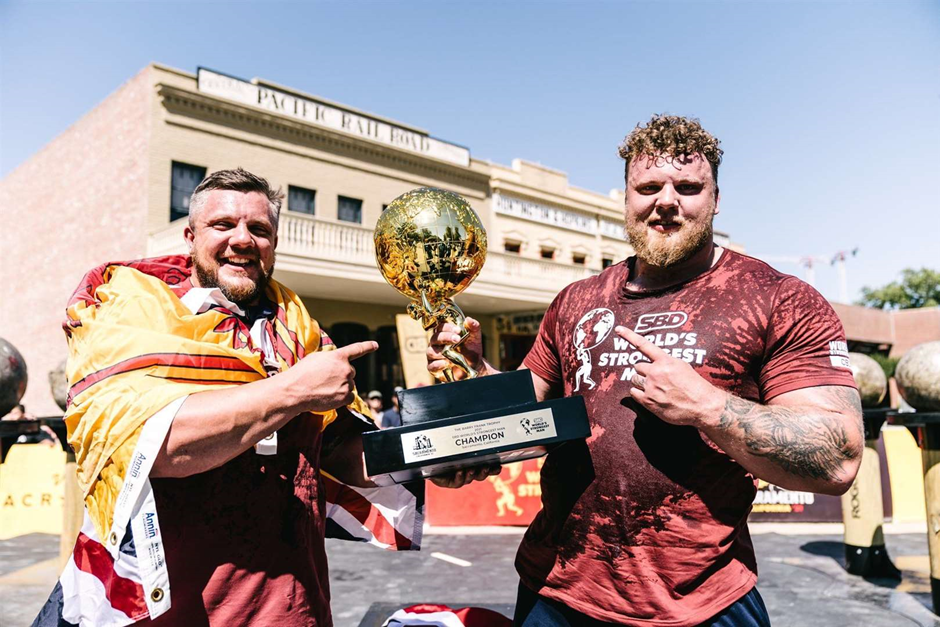 Tom and Luke Stoltman smiling and pointing at the trophy after winning the 2021 worlds strongest man competition.