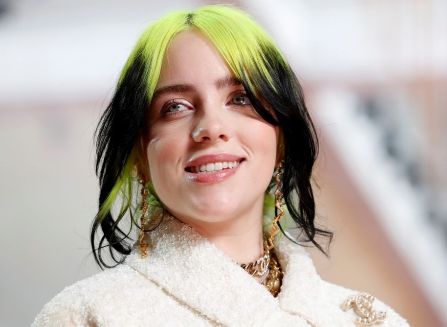 Billie Eilish with green and black shoulder length hair in a cream coat smiling.