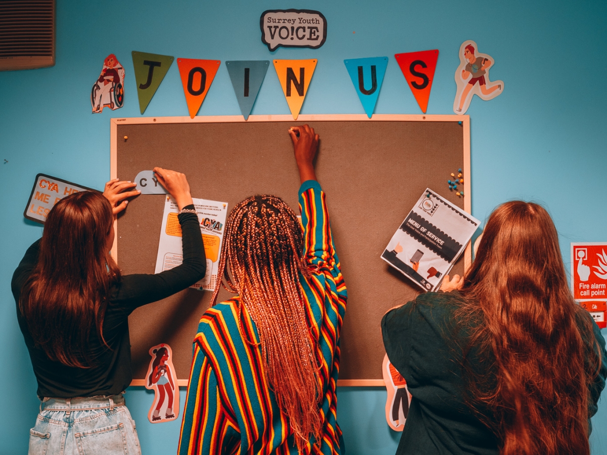 Three young people are decorating a notice board. Text reads Surrey Youth Voice. Join Us. There are cartoon images of young people around the board.