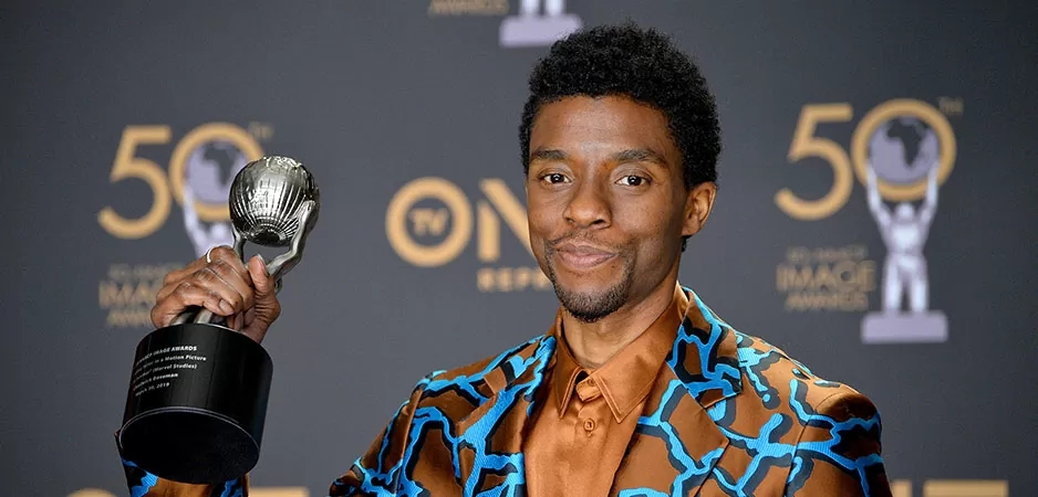 Chadwick is wearing a patterned light brown and light blue silk suit and light brown shirt. He is holding an award that he has won.
